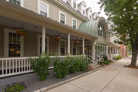 Exterior view of Zelma Lacey House showing a large porch with white railings and columns, hanging flower baskets, and a green arched roof over the entrance. There are shrubs and flowers along the building and a sidewalk with trees on the right side.