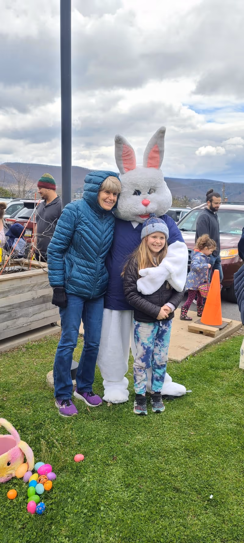 A woman and a young girl standing outside on grass, posing with a person dressed in a large white Easter Bunny costume. The woman is wearing a blue jacket and purple shoes, and the girl is wearing a black jacket, colorful pants, and a gray beanie. In the background, there are other people, parked cars, a wooden planter, and a cloudy sky with mountains in the distance. Colorful plastic Easter eggs are scattered on the grass in the foreground.