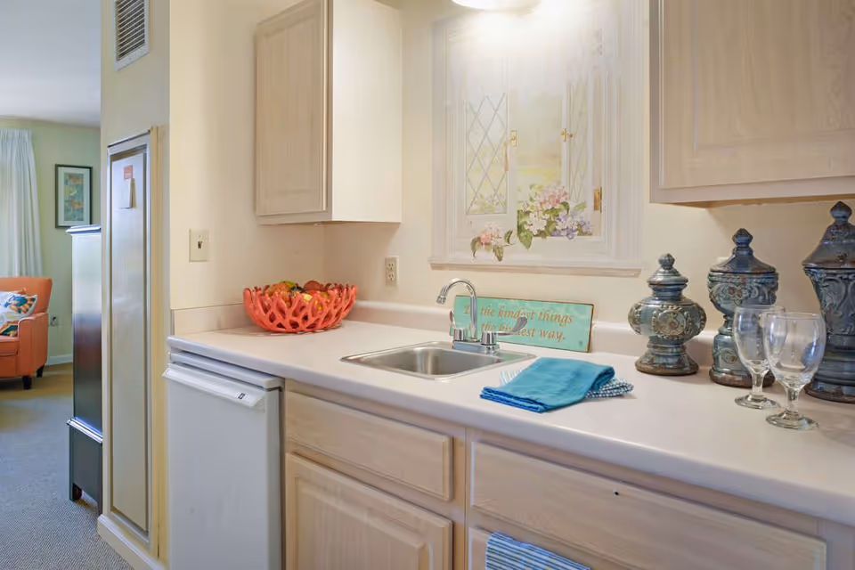 A small kitchen area with a countertop featuring a sink, a red fruit basket, decorative jars, and two empty wine glasses. A blue towel is folded next to the sink. In the background, there is a partial view of a living room with an orange armchair and framed artwork on the wall.