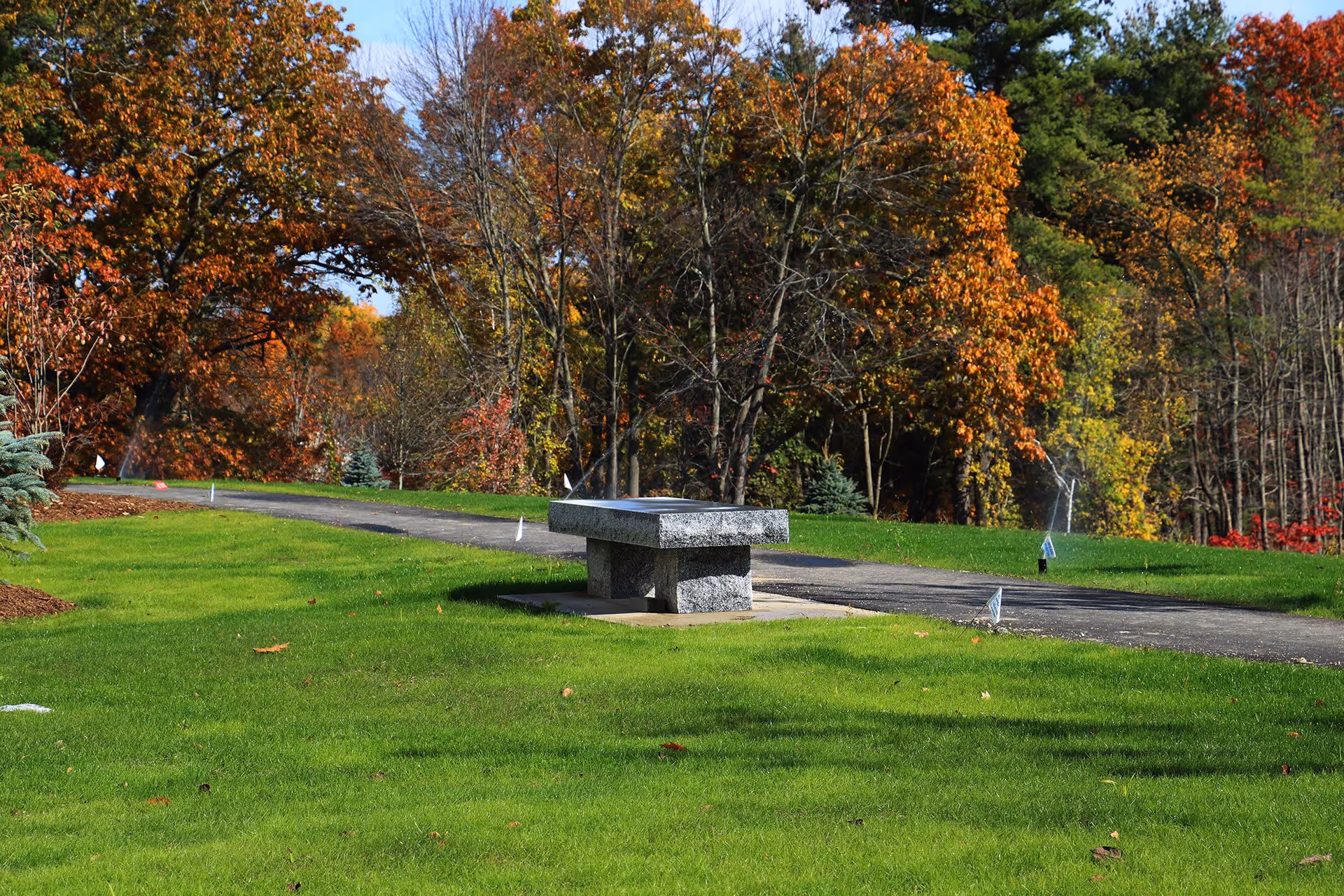 A stone bench sits on a concrete slab beside a paved walking path surrounded by green grass and trees with autumn foliage in shades of orange, yellow, and red.