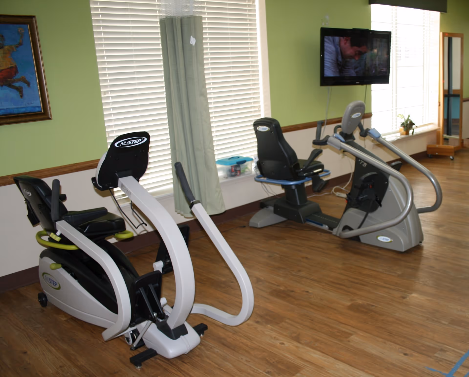 A rehab facility exercise room with two recumbent stationary bikes facing a wall-mounted TV.