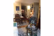 Interior view of a living and dining area featuring a wooden dining table with chairs, a patterned rug, a small round glass table with decorative items including candles, and a floral upholstered armchair. The room is warmly lit with lamps and decorated with framed artwork on the walls.
