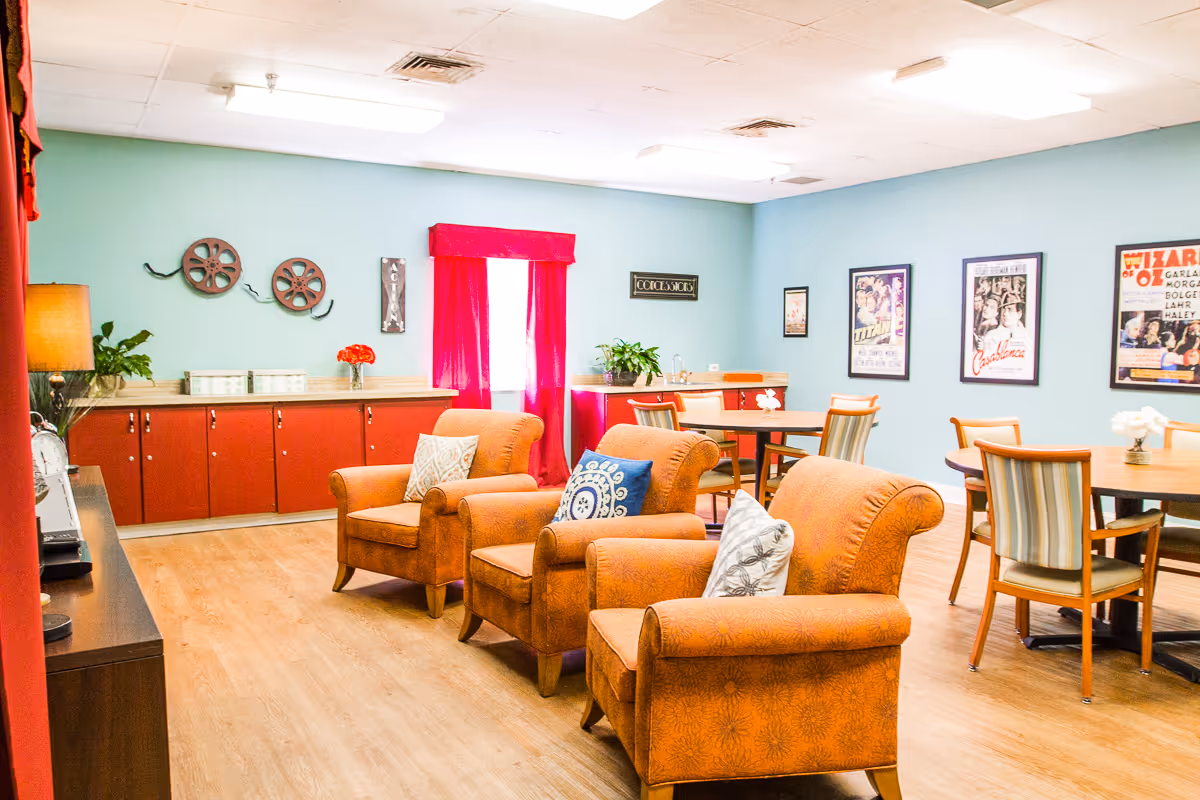 A bright and cozy common area in a senior living facility featuring three orange upholstered armchairs with decorative pillows arranged in front of a TV stand. Behind the chairs are round wooden tables with chairs, a red cabinet along the wall, and movie-themed wall art. The room has light blue walls, wood flooring, and a window with red curtains.