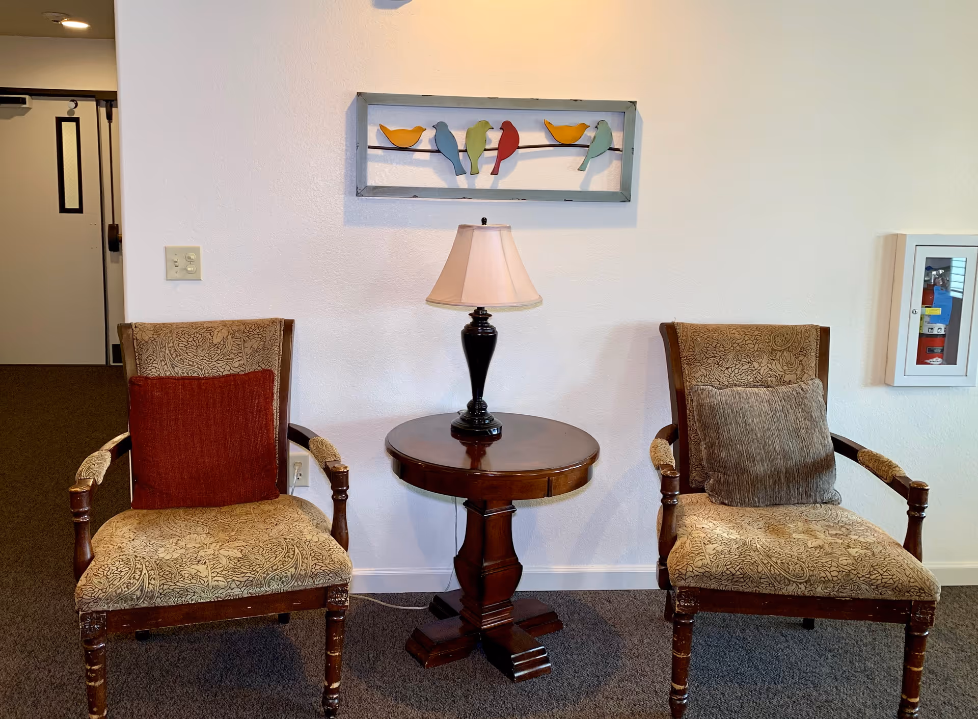 Two upholstered armchairs with patterned fabric and cushions, one red and one gray, are positioned on either side of a round wooden table with a black lamp on it. Above the table is a wall decoration featuring colorful bird figures on wires. The setting is indoors with a white wall and carpeted floor.