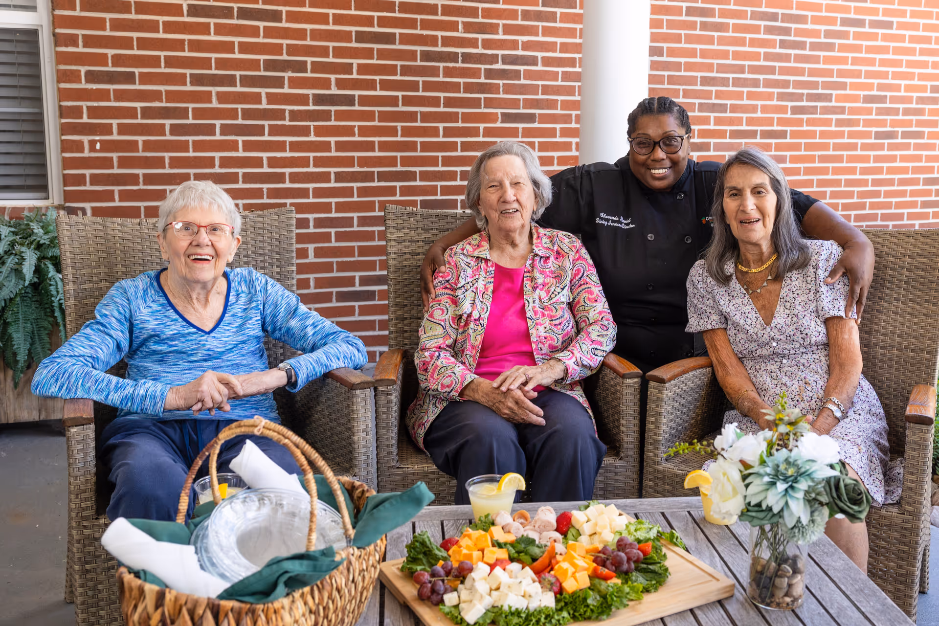Three elderly women sitting on wicker chairs on a patio with a smiling staff member standing behind them. A wooden table in front holds a platter of assorted cheeses, grapes, and rolled deli meats, along with two drinks garnished with lemon slices and a vase with white flowers. The background features a brick wall and a white column.