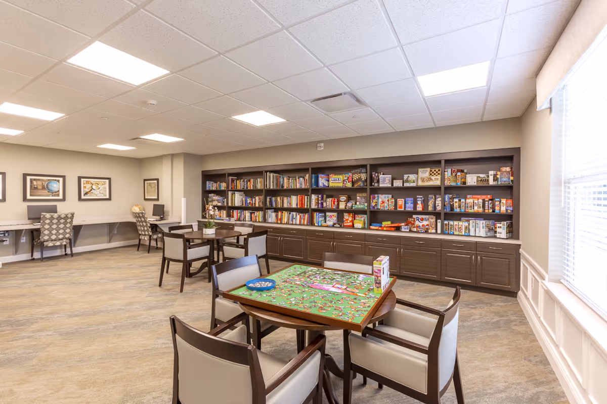 A well-lit activity room with tables and chairs arranged for games and puzzles. One table in the foreground has a partially completed jigsaw puzzle with puzzle pieces and a box on it. The back wall features large shelves filled with books and various board games. There are also two workstations with chairs and framed pictures on the wall to the left. A large window with blinds is on the right side of the room.