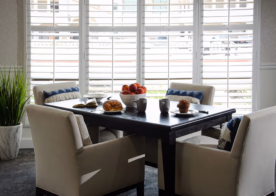 A dining area with a dark wooden table surrounded by four beige upholstered chairs, each with a blue and white decorative pillow. On the table are two coffee mugs, a croissant on a plate, a muffin on a plate, an open book, and a bowl of apples. Behind the table are large windows with white plantation shutters, and a potted green plant is visible to the left.
