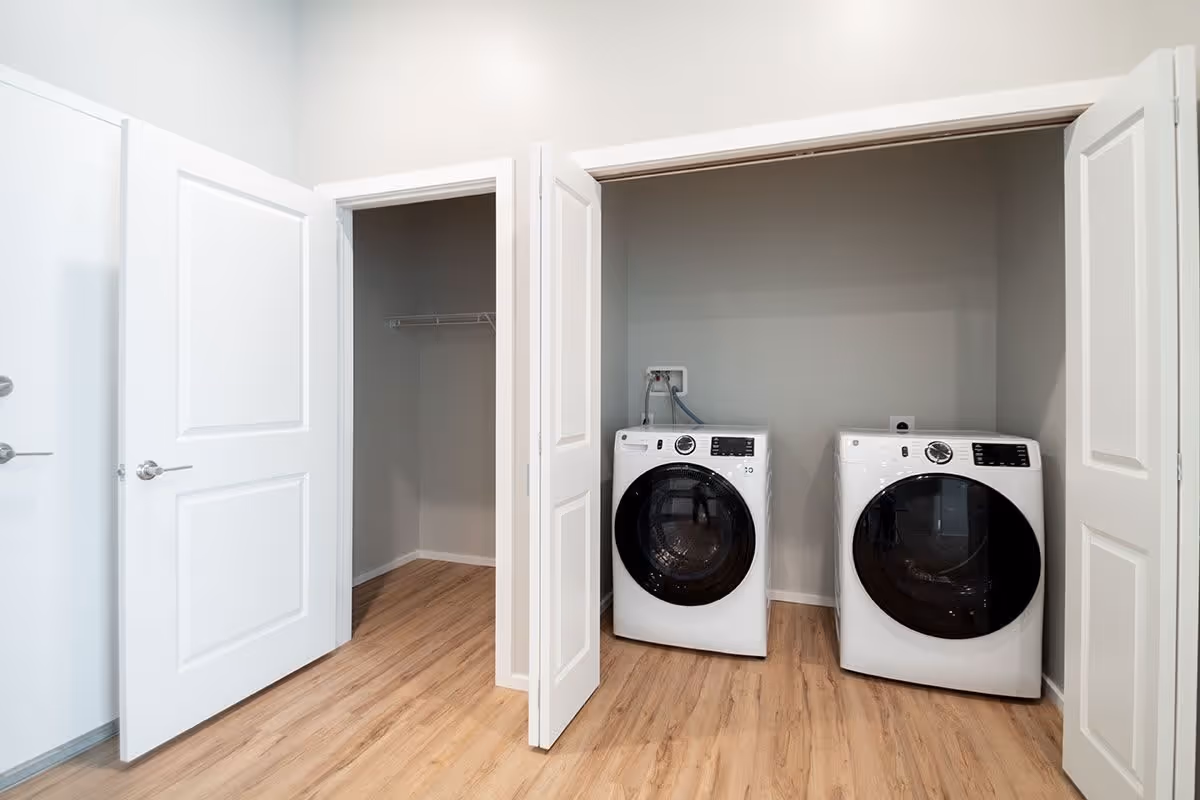 A laundry area with a white front-loading washing machine and dryer side by side inside a closet with white bi-fold doors. Next to the laundry closet is an open door revealing an empty closet with a hanging rod. The floor is light wood, and the walls are painted light gray.