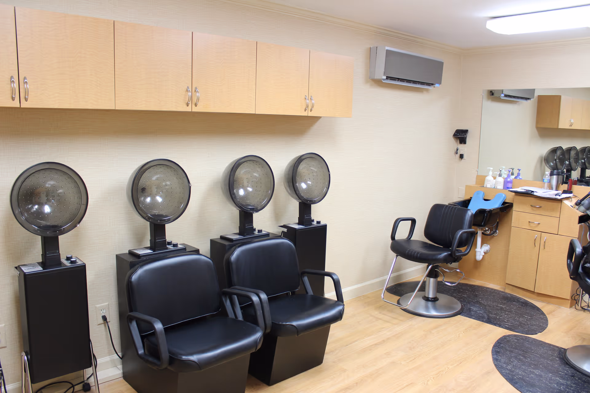 Interior view of a salon area with four black hair drying chairs with hooded dryers, a black styling chair, wooden cabinets, a large mirror, and various hair care products on the counter.