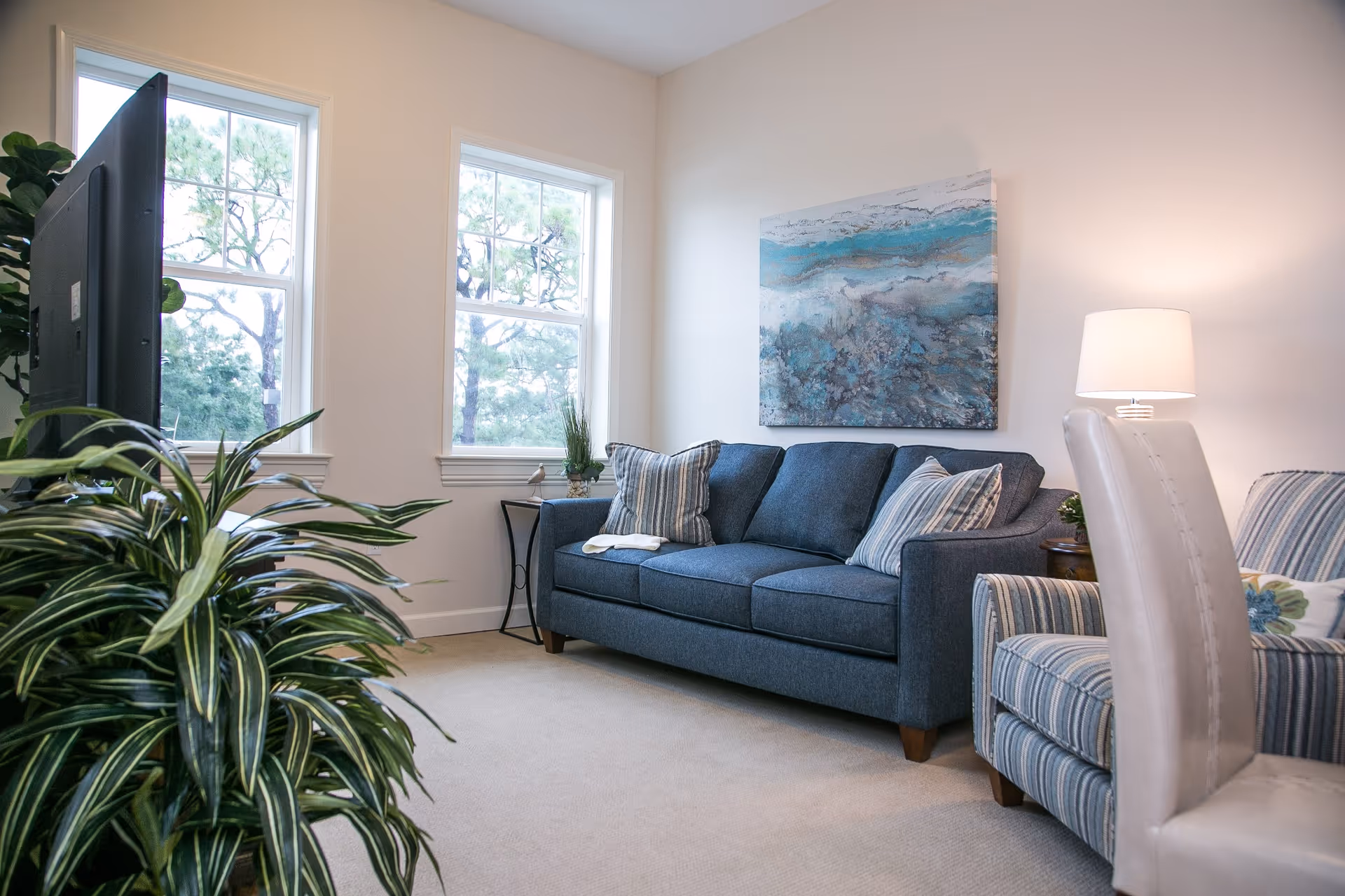 A cozy living room with a blue sofa adorned with striped pillows, a striped armchair, a white leather chair, a side table with a lamp, and two windows letting in natural light. A large abstract painting with blue and gray tones hangs on the wall above the sofa. There are green plants near the television and by the window.