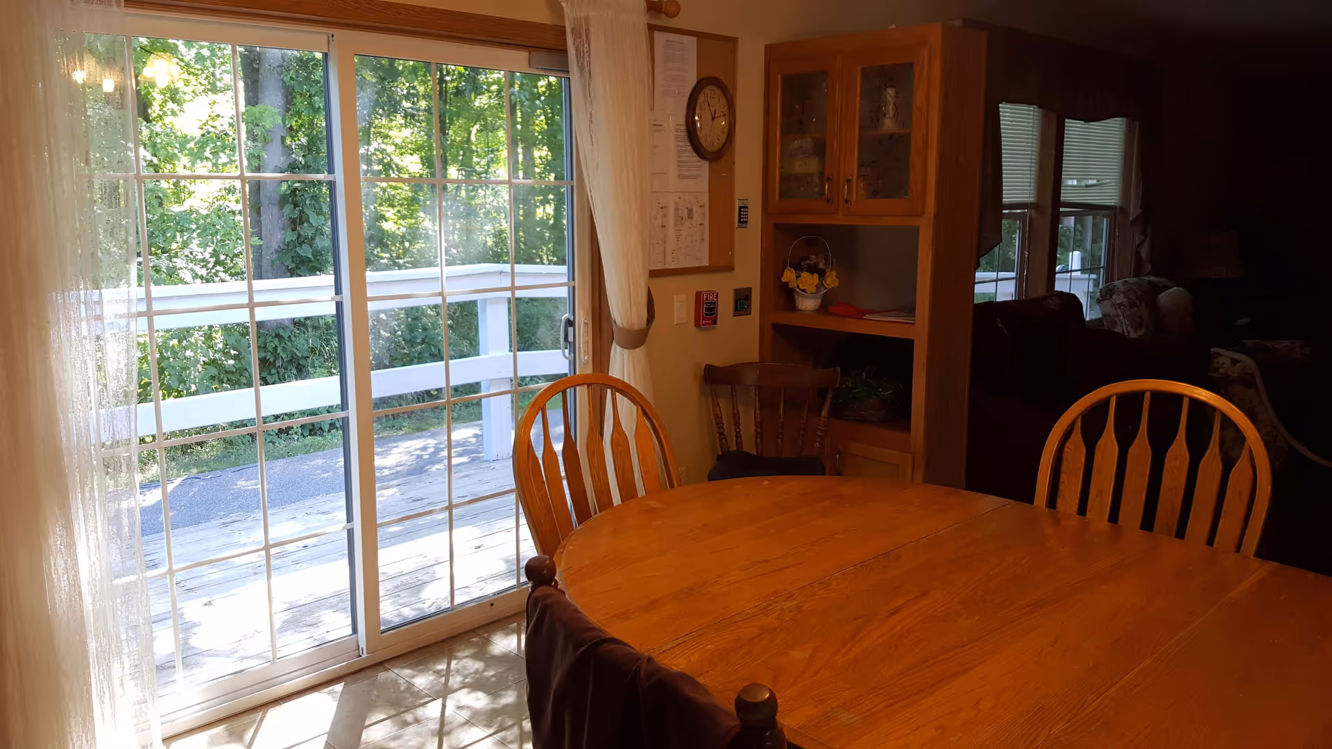 A wooden dining table with chairs is positioned near a large sliding glass door that opens to an outdoor deck with a white railing and green trees beyond. Inside, there is a wooden cabinet with glass doors, a wall clock, and a bulletin board with papers pinned to it. The room is softly lit with natural light coming through the glass door and windows.
