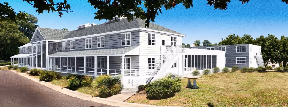 Front exterior of a two-story white and gray building with a wraparound porch, lawn, and driveway under a blue sky.