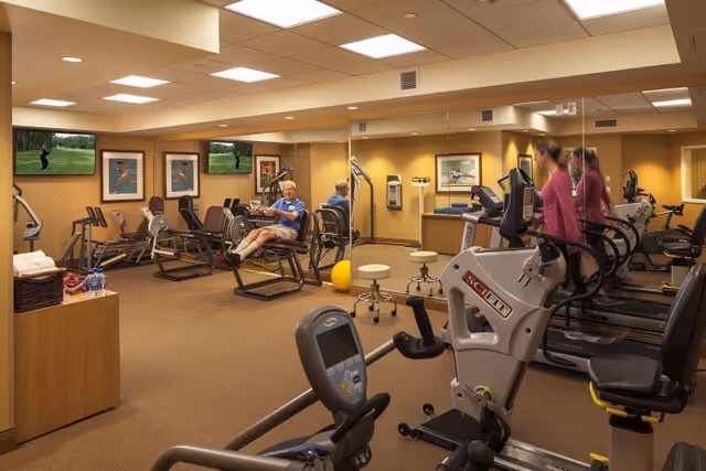 An indoor fitness room with treadmills, recumbent bikes, and other exercise equipment where seniors are exercising.