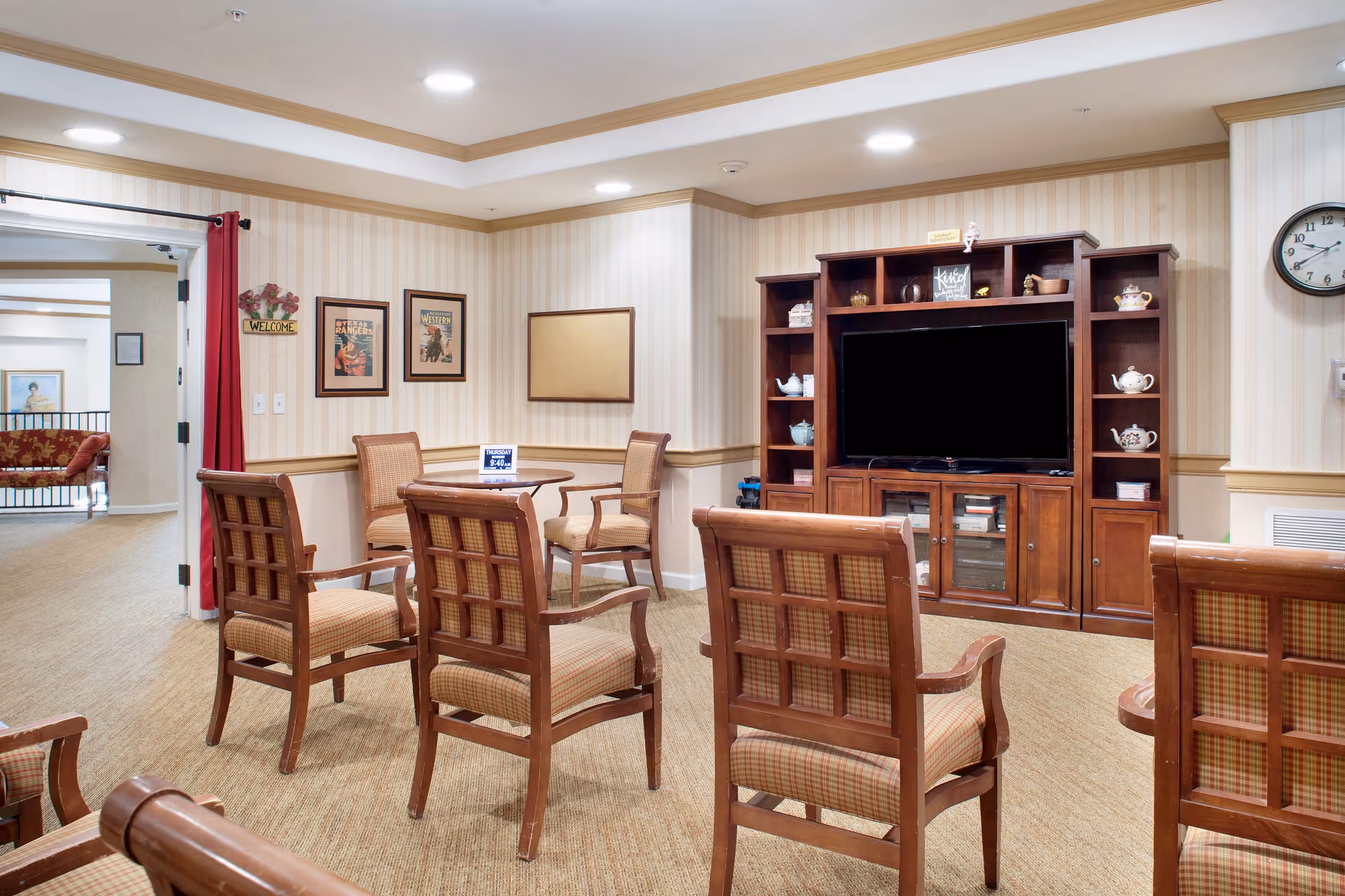 A cozy common area with several wooden chairs arranged facing a large wooden entertainment center with a flat-screen TV. The walls have light-colored striped wallpaper with framed pictures and a clock. There is a small round table with chairs in the corner and an open doorway with a red curtain leading to another room.