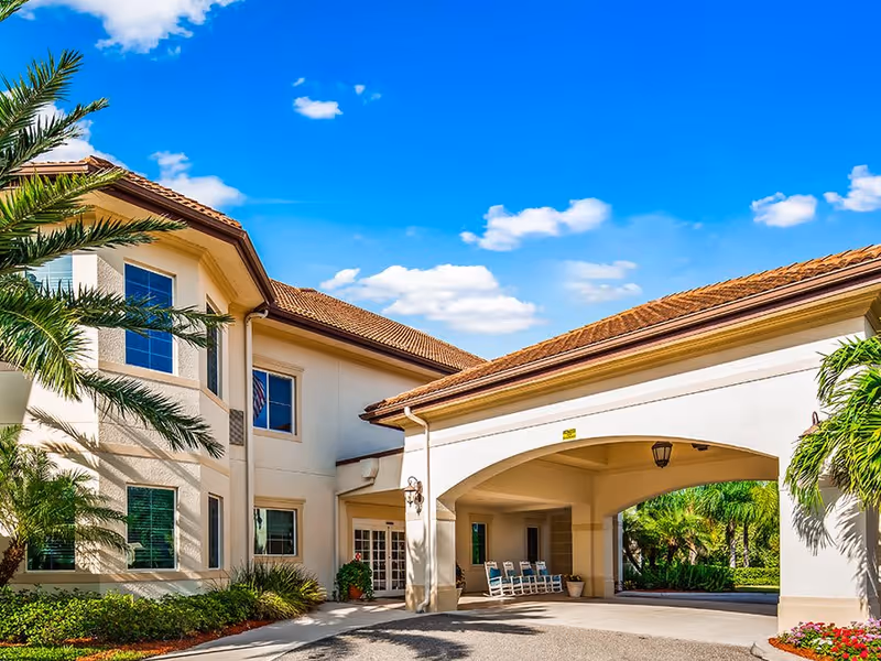 Exterior view of The Windsor of Venice senior living facility showing a two-story building with beige walls and a red-tiled roof under a bright blue sky with some clouds. There are palm trees and landscaped greenery around the entrance, which features a covered driveway with rocking chairs.