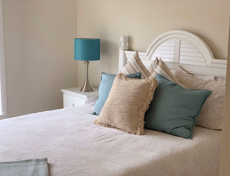 A bedroom featuring a white bed with layered throw pillows, a white nightstand and a teal table lamp.
