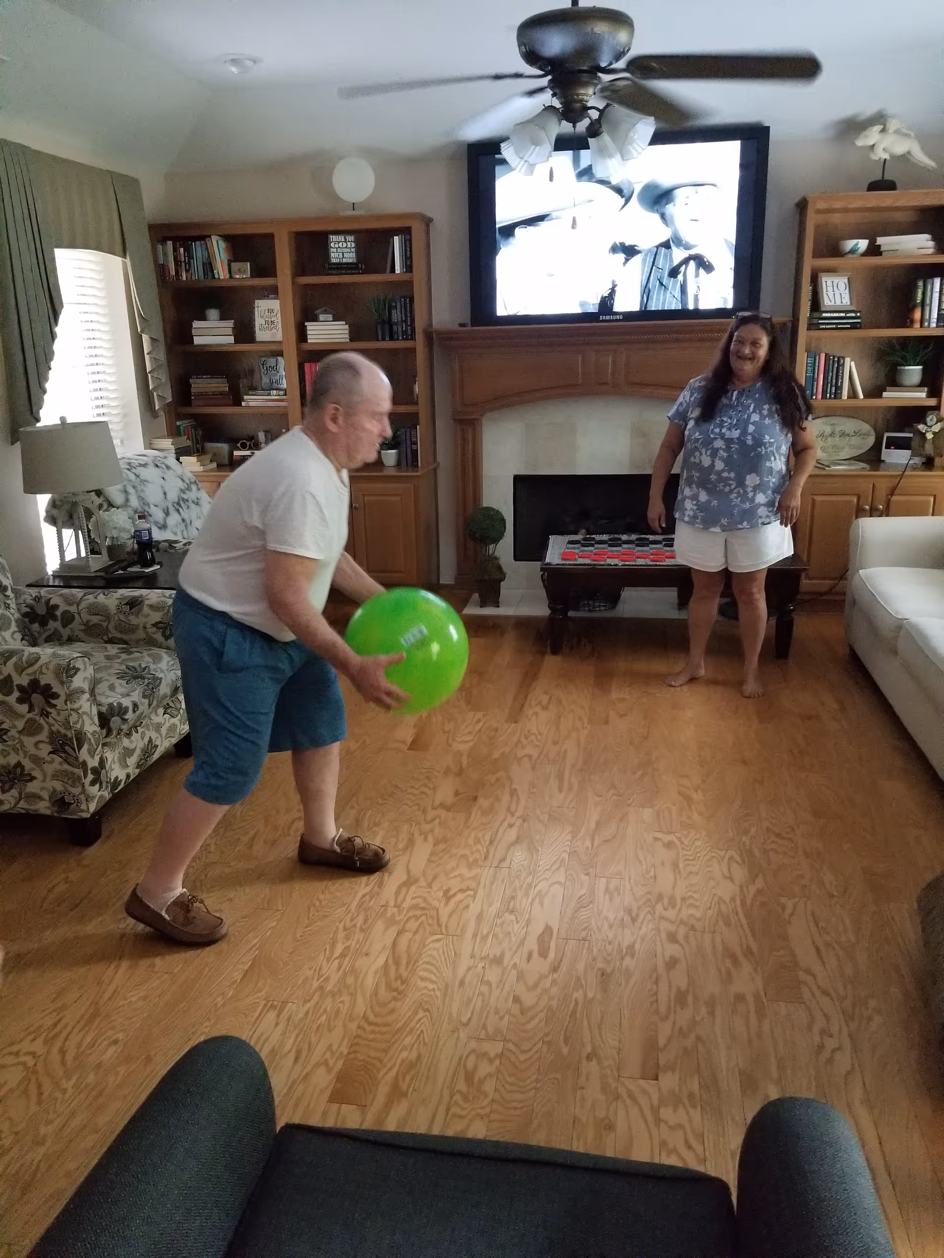 Two people playing with a green ball in a living room with hardwood floors, a TV above a fireplace, and bookshelves.