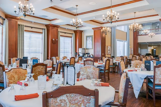 A spacious dining room with multiple round tables covered with white tablecloths, each set with glasses, napkins, and menus. The room features wooden columns, large windows with blinds, and elegant chandeliers hanging from the ceiling. Upholstered chairs with floral patterns surround the tables.