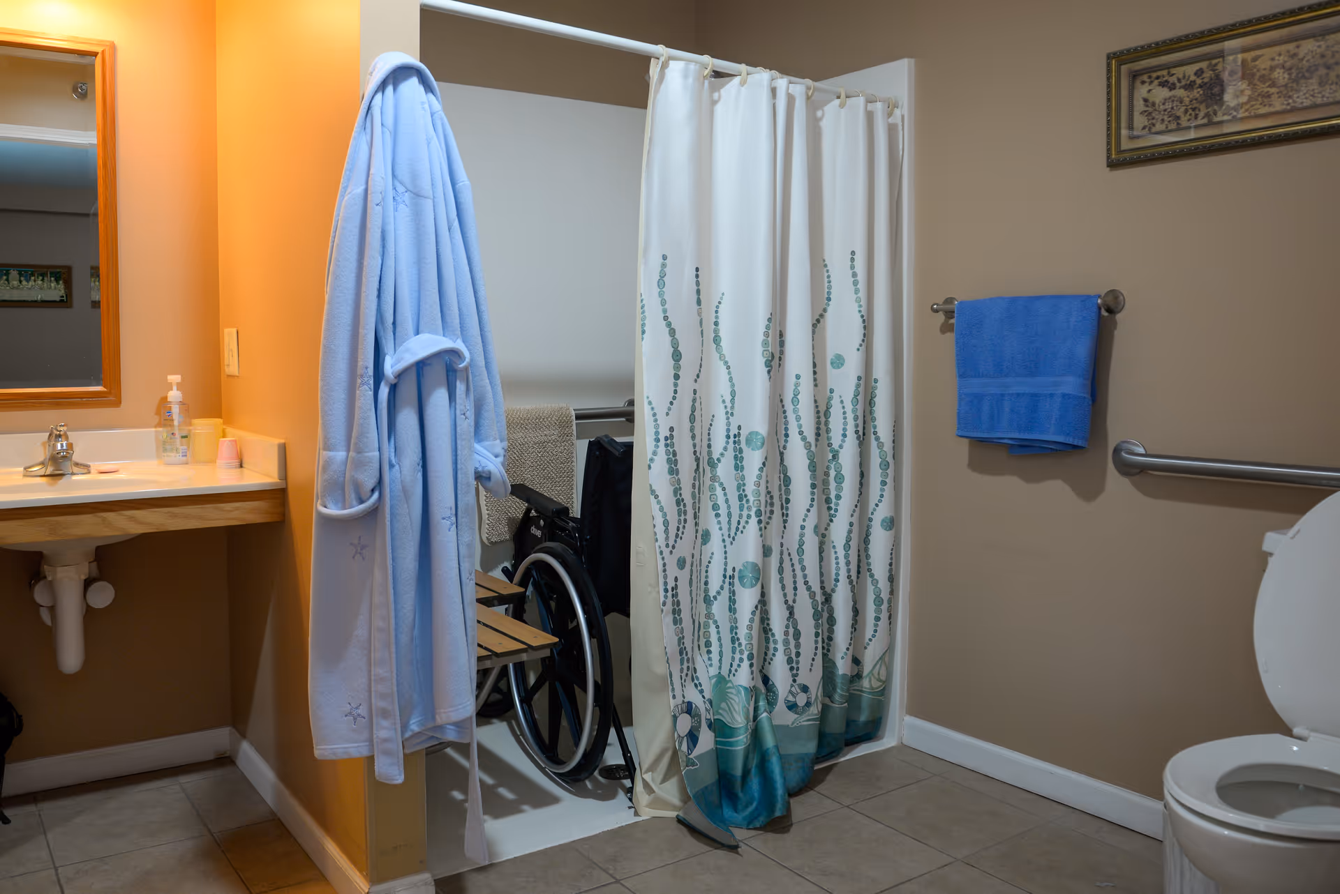 Accessible bathroom with a shower curtain partially open revealing a wheelchair, a sink with mirror, toilet, grab bar, and a blue robe and towel.