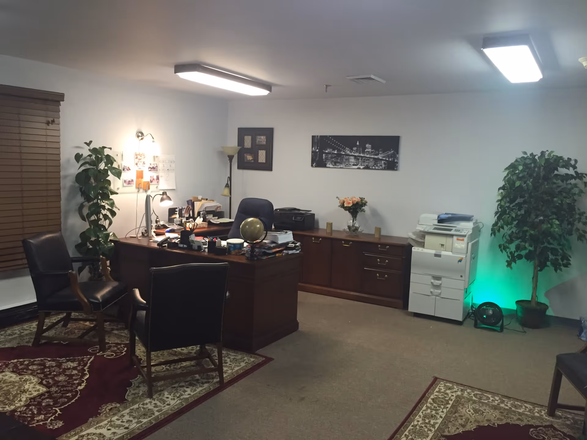 An office room with a wooden desk cluttered with papers, a globe, and office supplies. Two black leather chairs are positioned in front of the desk. The room has beige carpet with patterned rugs, a large wooden cabinet along the back wall, a printer, and two potted plants. The walls are white with framed pictures and a black and white cityscape photo. The room is lit by ceiling lights and a wall lamp.