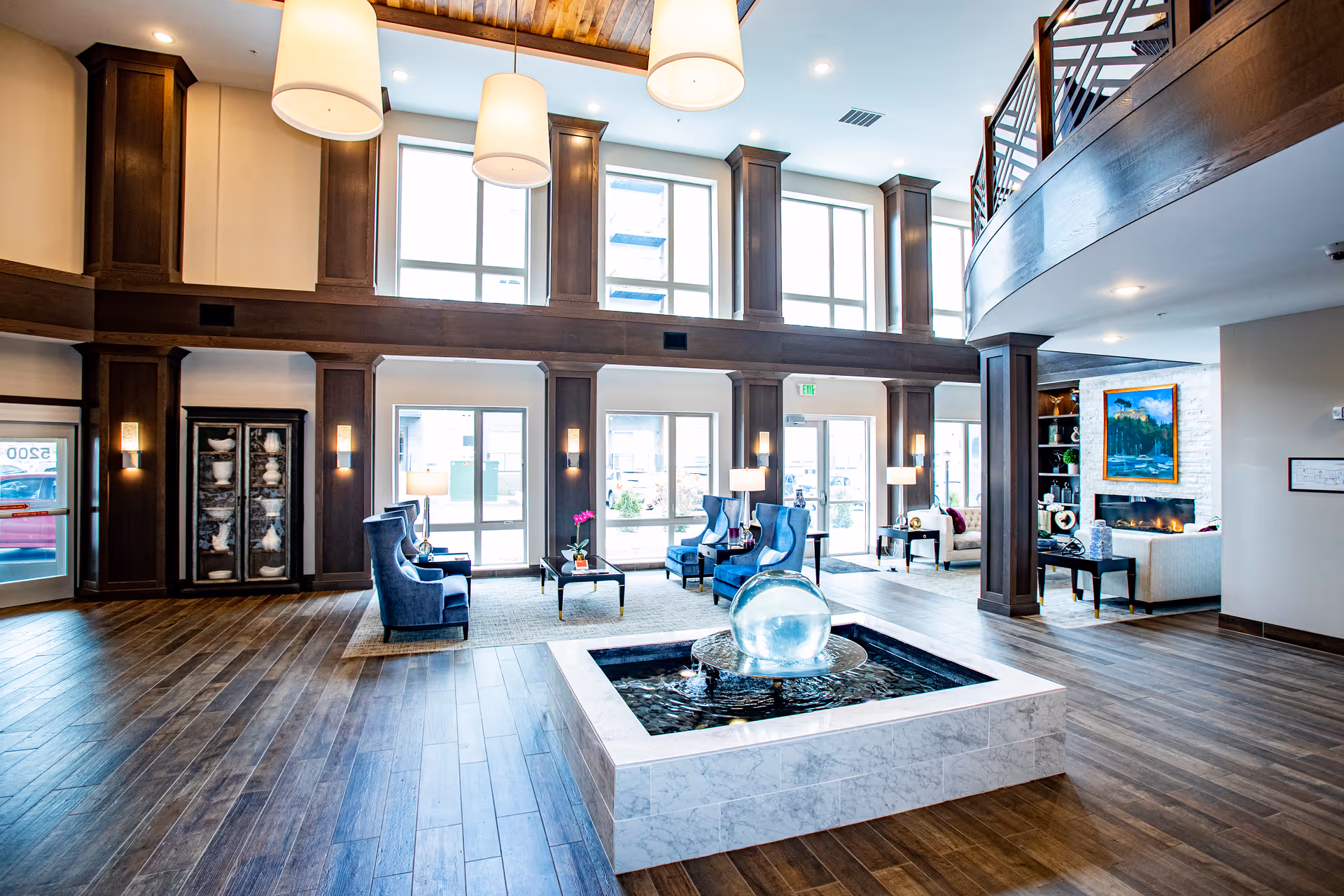Spacious and bright senior living facility lobby with high ceilings, large windows, wooden flooring, and a modern water fountain in the center. The seating area includes blue armchairs and coffee tables, with additional seating and a fireplace visible in the background.