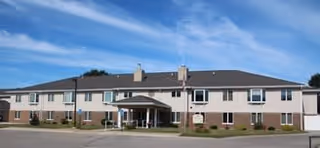 Exterior view of a two-story senior living facility building under a clear blue sky, featuring multiple windows and a covered entrance with a driveway in front.