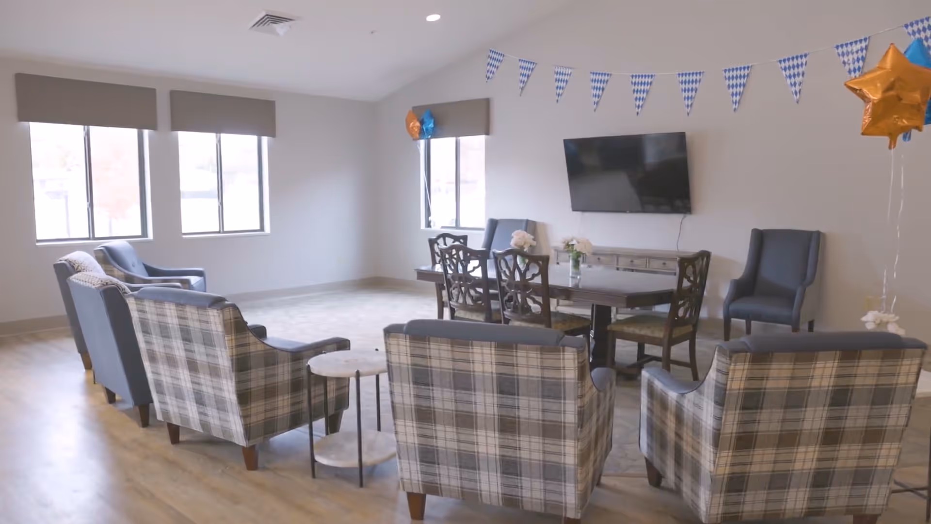 A bright and spacious common area with multiple plaid armchairs arranged around small round tables. There is a wooden dining table with chairs against the far wall, a flat-screen TV mounted above a console table, and festive blue and white pennant banners along with star-shaped balloons decorating the room. Large windows allow natural light to fill the space.