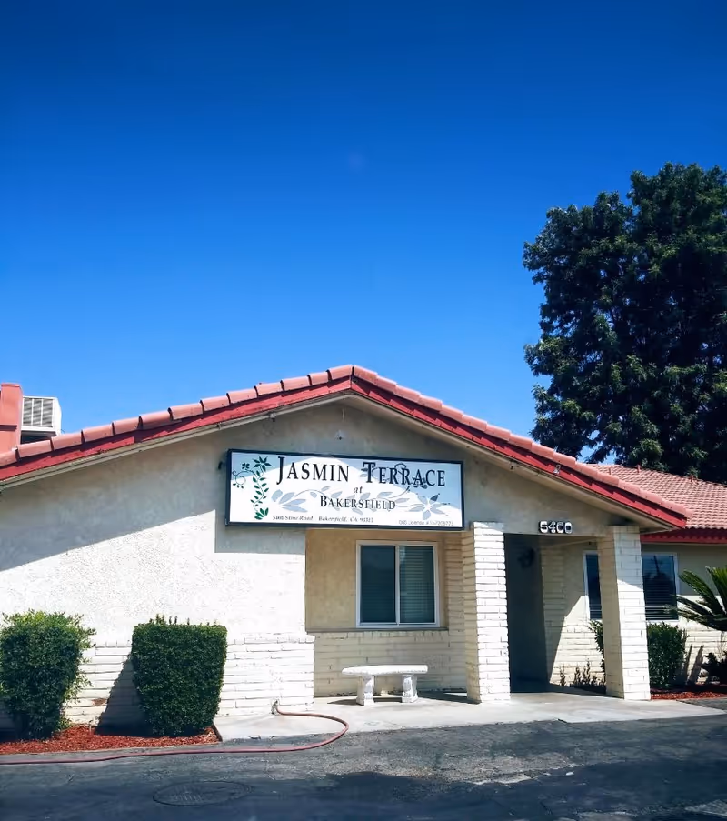 Exterior view of Jasmin Terrace at Bakersfield, a single-story building with a red tiled roof, beige walls, a small bench in front, and some bushes along the building. The sky is clear and blue.
