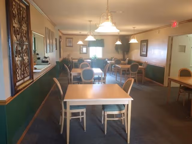 A dining room with several wooden tables and chairs arranged neatly. The walls are painted green on the lower half and beige on the upper half, decorated with framed pictures and a decorative metal wall hanging. Ceiling lights hang above the tables, and a window at the far end lets in natural light.