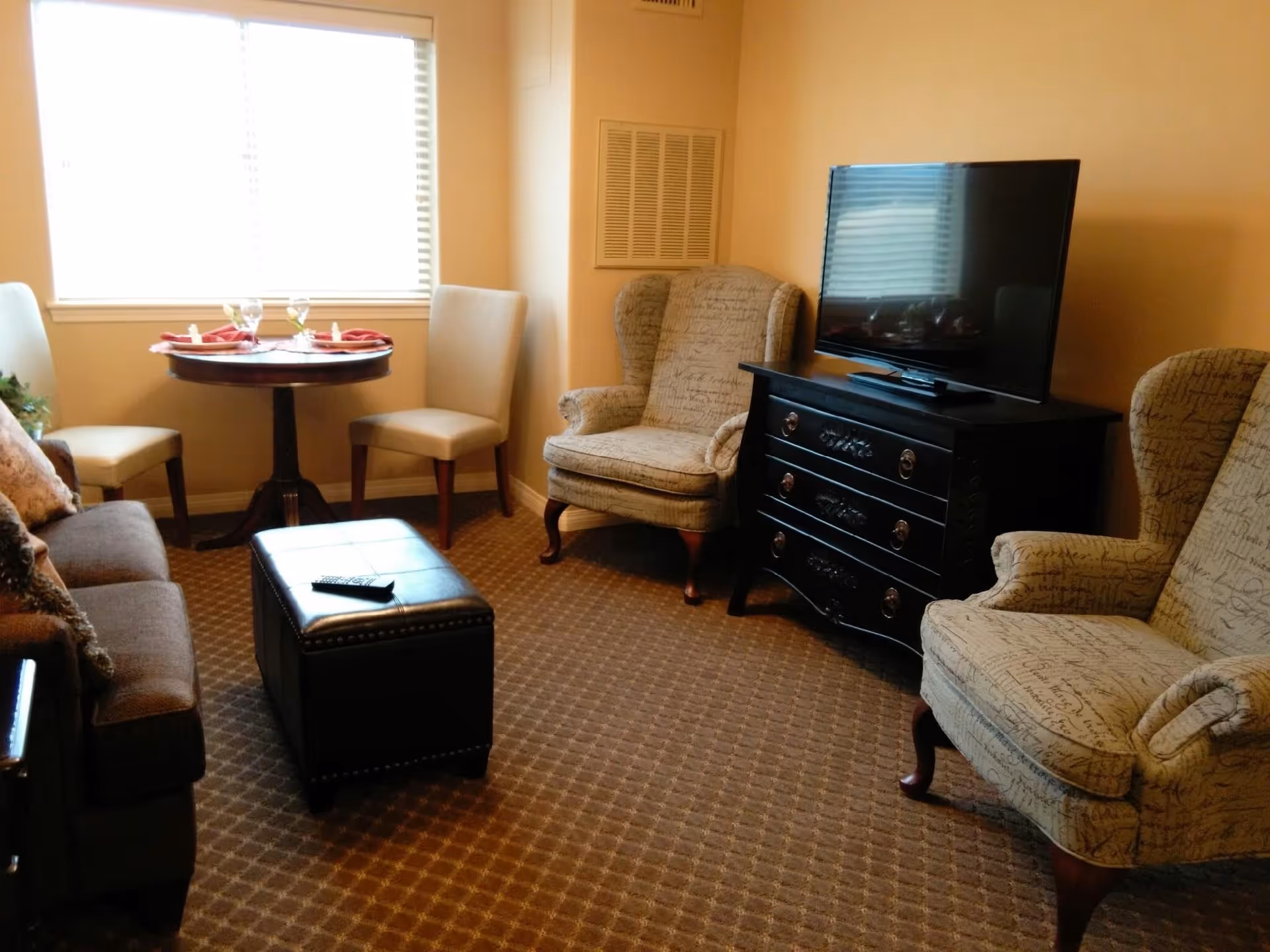 A cozy living room with two patterned armchairs, a dark wooden dresser with a flat-screen TV on top, a small round table set with two glasses and napkins, two beige chairs, a brown sofa with cushions, and a black ottoman with a remote control on it. The room has a window with blinds letting in natural light and a carpeted floor.
