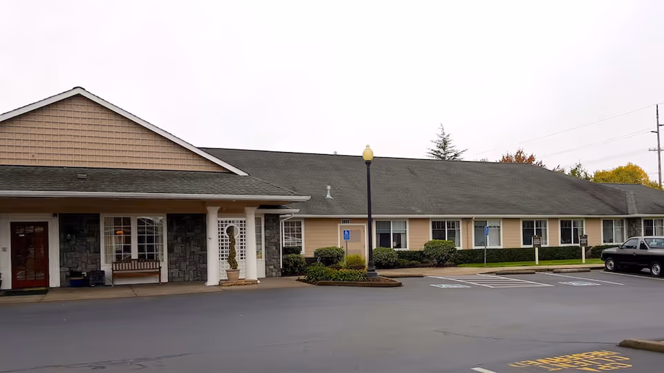 Exterior view of a single-story building with beige siding and a dark gray roof, featuring a covered entrance supported by white columns, a bench near the entrance, several windows, and a parking lot with marked handicap spaces and a black pickup truck parked on the right side.
