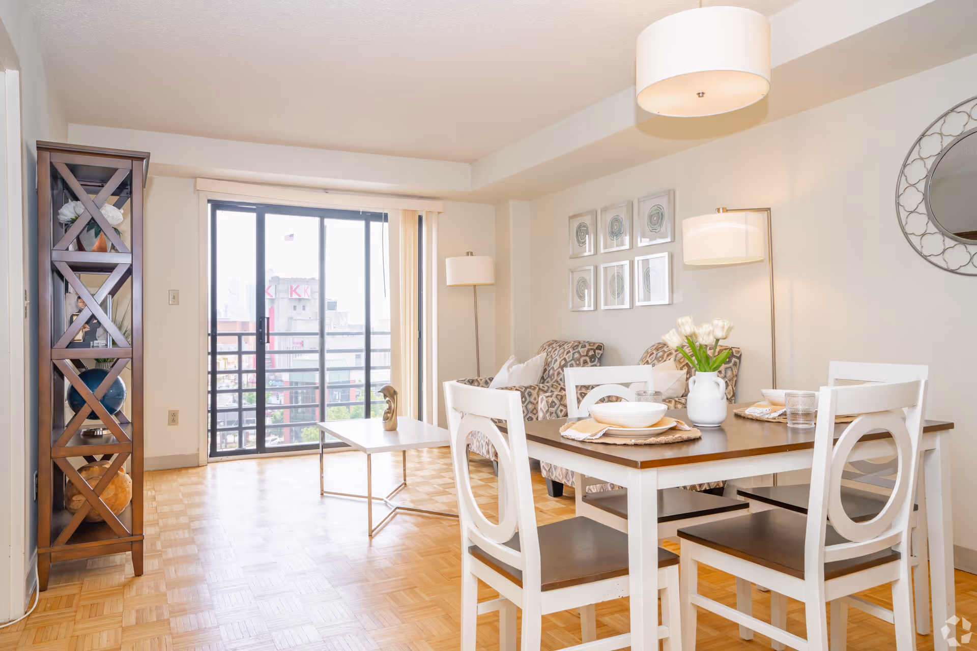 Bright and airy living and dining area with a wooden dining table set for four, two patterned armchairs, a coffee table, a tall wooden shelving unit with decorative items, and large sliding glass doors leading to a balcony with a city view.