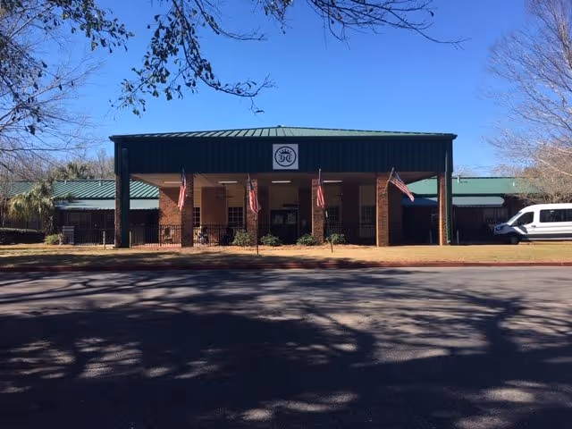 Front exterior view of Crowne Health Care of Mobile building with a green roof, brick pillars, and four American flags displayed in front. There is a white van parked on the right side and leafless trees framing the scene under a clear blue sky.