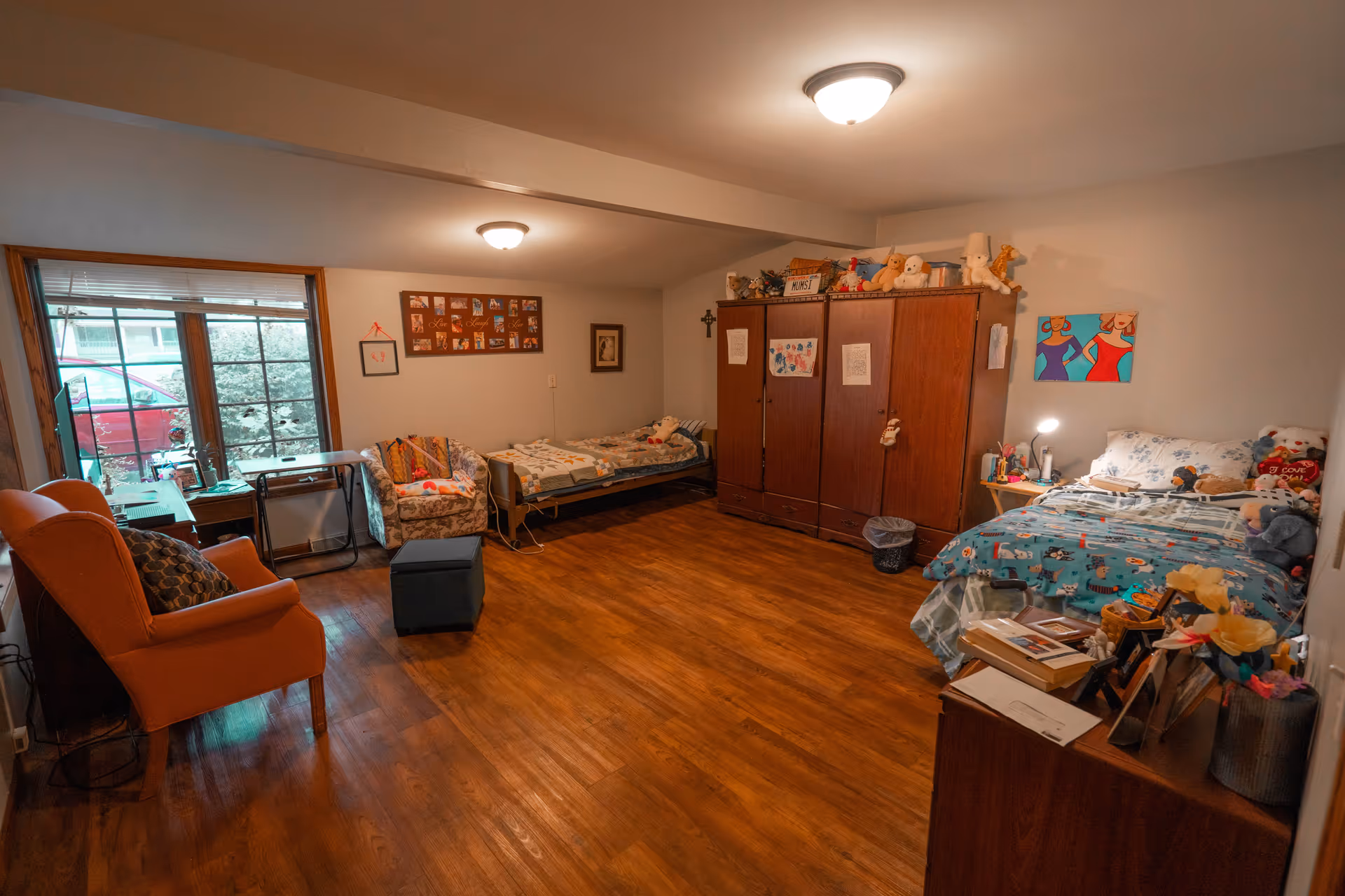 A cozy room with two beds, a wooden wardrobe, a floral armchair, an orange armchair, and a small table with a lamp. The room has wooden flooring and light-colored walls decorated with pictures and stuffed animals on the furniture. A large window shows a red car and greenery outside.