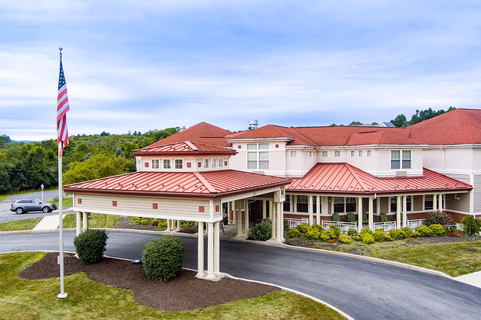Exterior view of a senior living facility with a covered entrance supported by columns, red metal roofs, beige siding, and an American flag on a flagpole in the front lawn. There are shrubs and landscaped areas around the building and a car parked in the driveway.