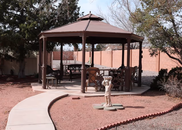 Outdoor gazebo with a brown roof and several chairs and tables underneath, surrounded by trees and a brick wall. A small statue of a child holding a basket on their head is placed on a stepping stone path in front of the gazebo. The ground is covered with reddish gravel and a curved concrete walkway leads to the gazebo.
