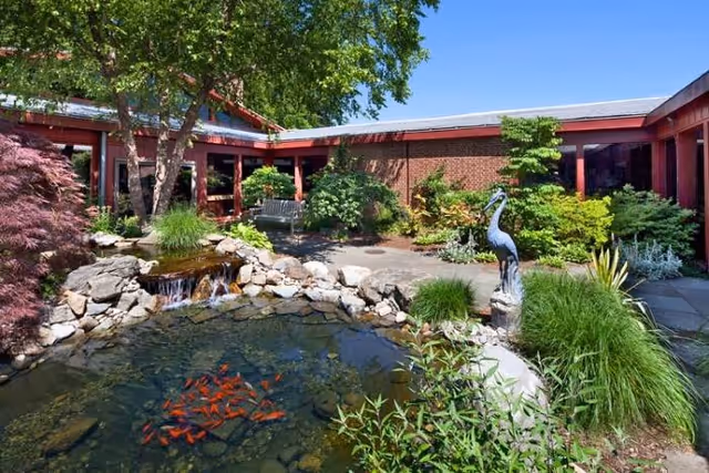 A peaceful outdoor courtyard at Meadow Lakes featuring a small pond with orange fish, a waterfall flowing over rocks, lush greenery, a metal bird sculpture, and a bench surrounded by trees and shrubs under a clear blue sky.