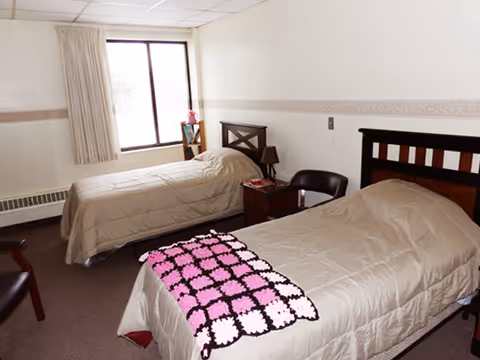 Simple assisted-living bedroom with two twin beds, a window, a nightstand, and chairs.