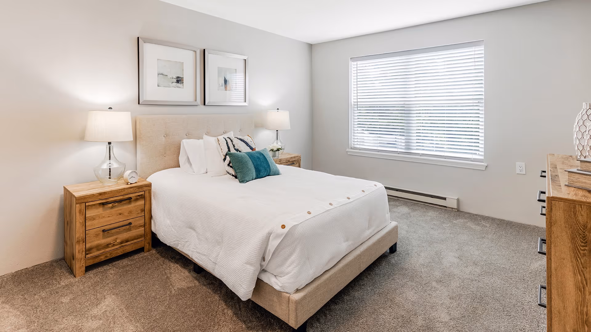 A bright and clean bedroom with a beige upholstered bed, white bedding, and decorative pillows. There are two wooden nightstands with lamps on either side of the bed, a window with white blinds, and a wooden dresser with decorative items on the right side. The room has light-colored walls and carpeted flooring.