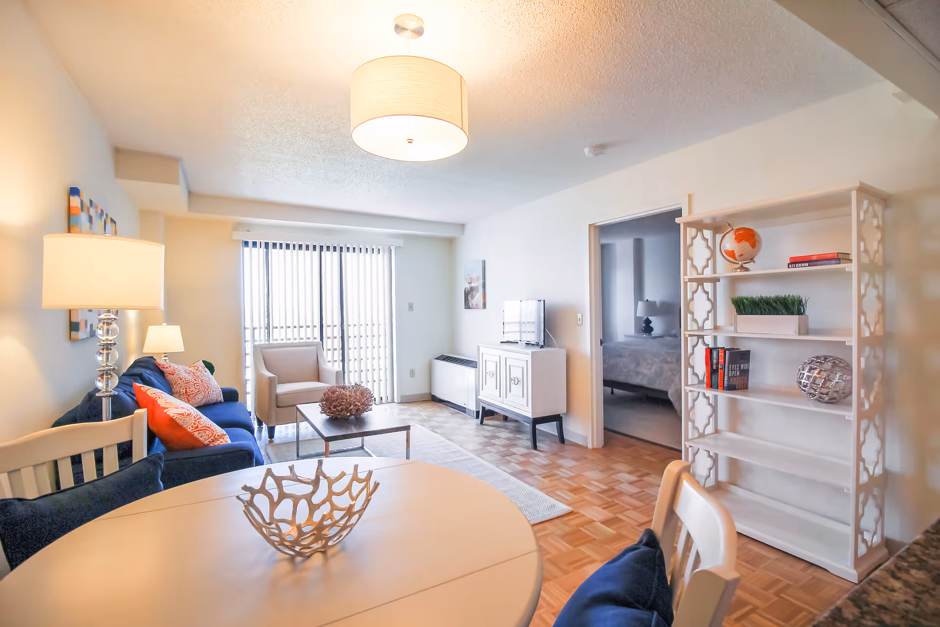 A bright and cozy living room in a senior living facility featuring a blue sofa with colorful pillows, a beige armchair, a coffee table with decorative items, a white round dining table with chairs, a white bookshelf with books and decorative objects, a small TV on a white cabinet, and a doorway leading to a bedroom. The room has parquet flooring and vertical blinds covering a large window or sliding door.