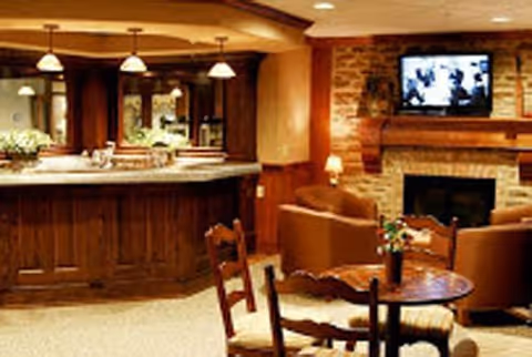 Cozy interior of a senior living facility common area featuring a wooden bar counter with pendant lights above, a stone fireplace with a mounted flat-screen TV, comfortable armchairs, and a round wooden table with chairs.