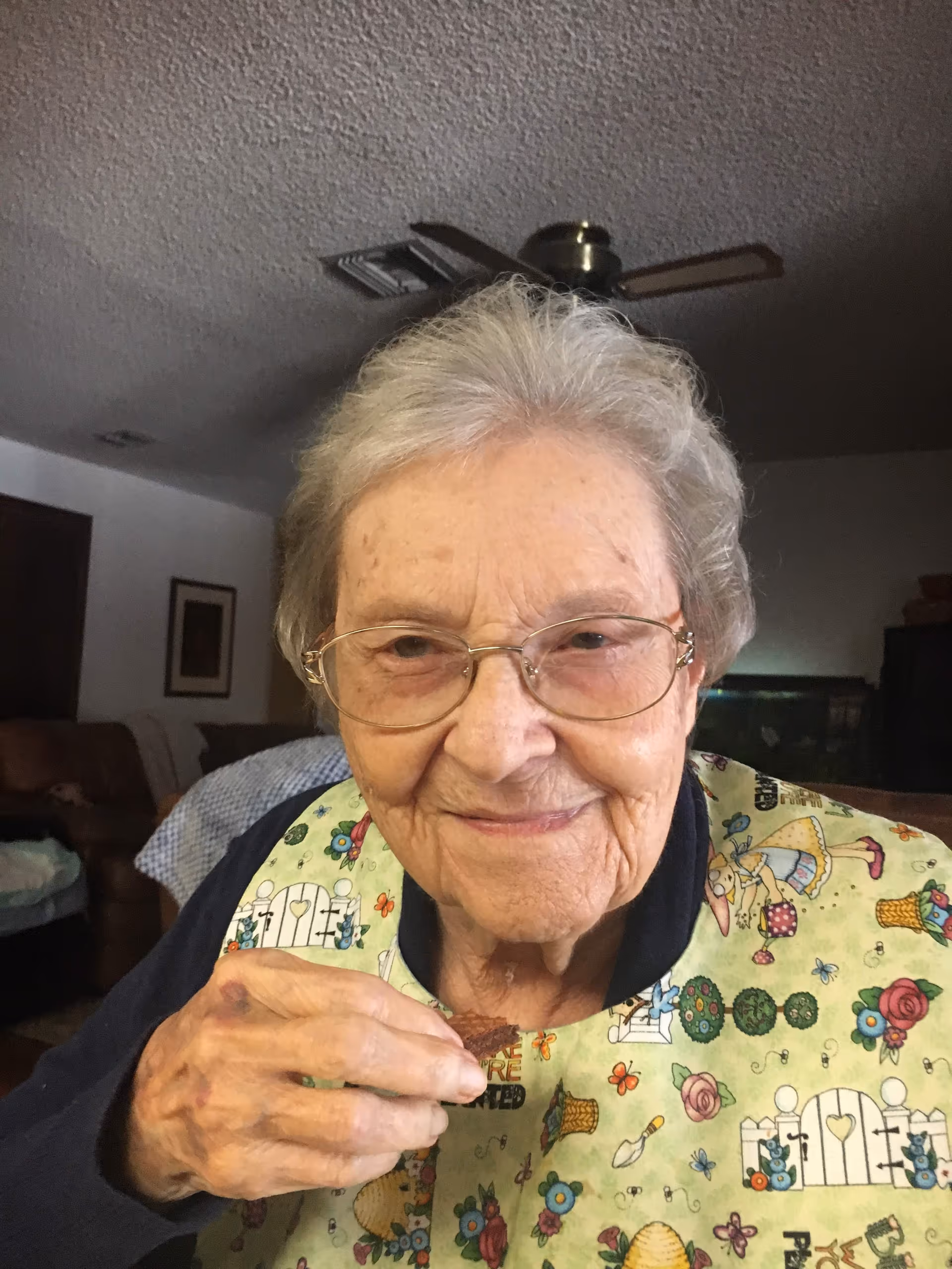 An elderly woman with glasses and gray hair is smiling at the camera while holding a piece of food. She is wearing a colorful bib with garden-themed illustrations. The background shows a living room with a ceiling fan, a framed picture on the wall, and some furniture.