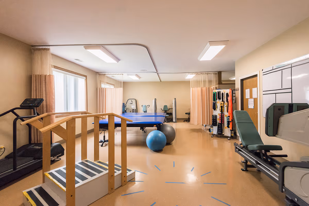 A senior living facility therapy/rehab room with exercise equipment including a treadmill, treatment table, therapy stairs, exercise balls and a weight machine.