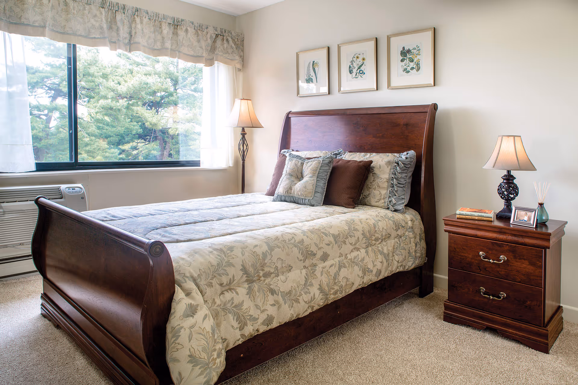 A bedroom with a wooden sleigh bed covered with a floral patterned comforter and multiple pillows. There is a wooden nightstand with a lamp, books, a framed photo, and a reed diffuser. A large window with sheer curtains and a valance shows green trees outside. The room has beige walls and carpeted floor.
