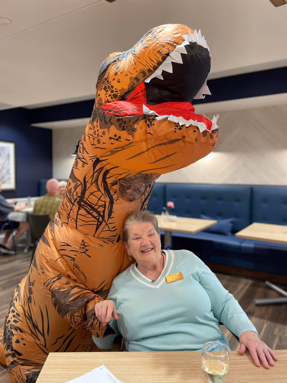 An elderly woman smiles at a dining table while someone in an inflatable T-Rex costume leans over her in a dining room.