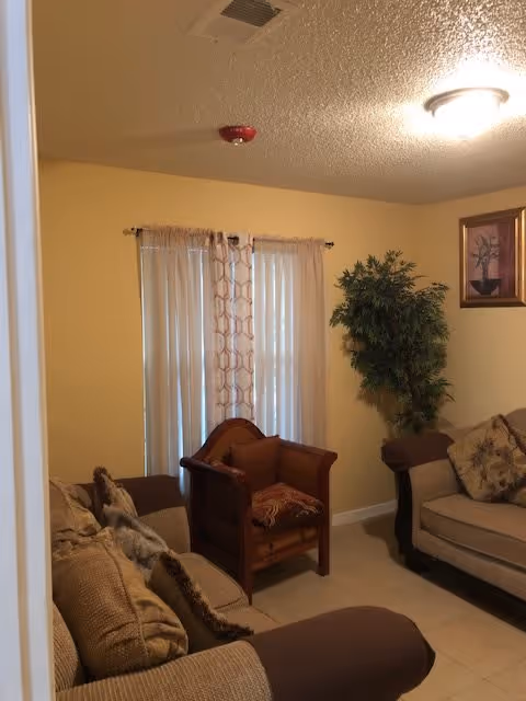 A cozy living room with beige walls and tiled floor, featuring two beige sofas with cushions, a wooden armchair with a patterned cushion, a tall green potted plant in the corner, a framed picture on the wall, and a window covered with sheer curtains and patterned drapes.