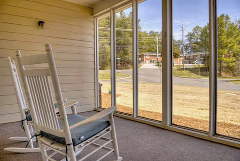 Two white wooden rocking chairs with cushions on a carpeted screened-in porch overlooking a grassy area and a road with trees and buildings in the background.