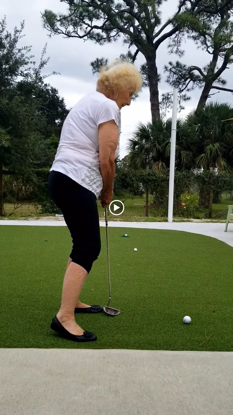 An elderly woman playing mini golf on a putting green outdoors, surrounded by trees and greenery.