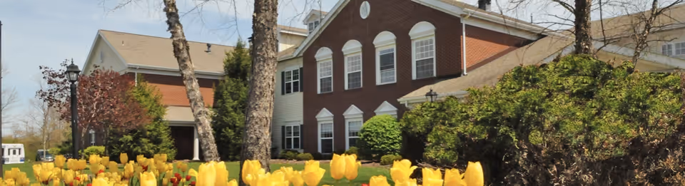 Exterior view of a brick and white senior living facility building surrounded by trees and bushes, with a garden of yellow tulips in the foreground under a clear sky.