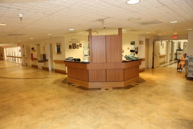 Interior view of a healthcare facility hallway with a central wooden nurse's station. The hallway has beige walls, a tiled ceiling with recessed lighting, and a light brown floor. There are doors and handrails along the walls, and a person in a wheelchair is visible in the background on the right side.